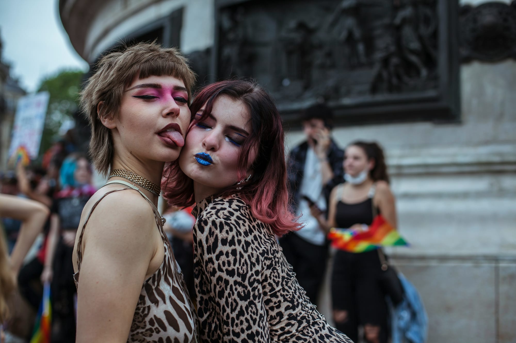 An image of two femme-presenting people, one with pink eye shadow sticking out their tongue, and one with blue lipstick, both wearing leopard-skin style clothing, in front of a small crowd.