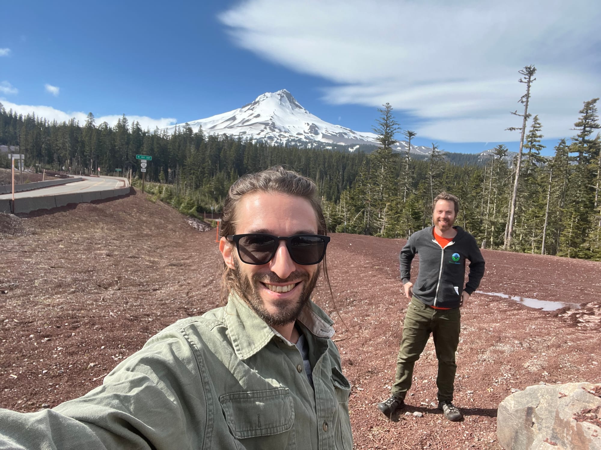Two guys in warm clothes standing on a gravel roadside in front of a snow-covered mountain.